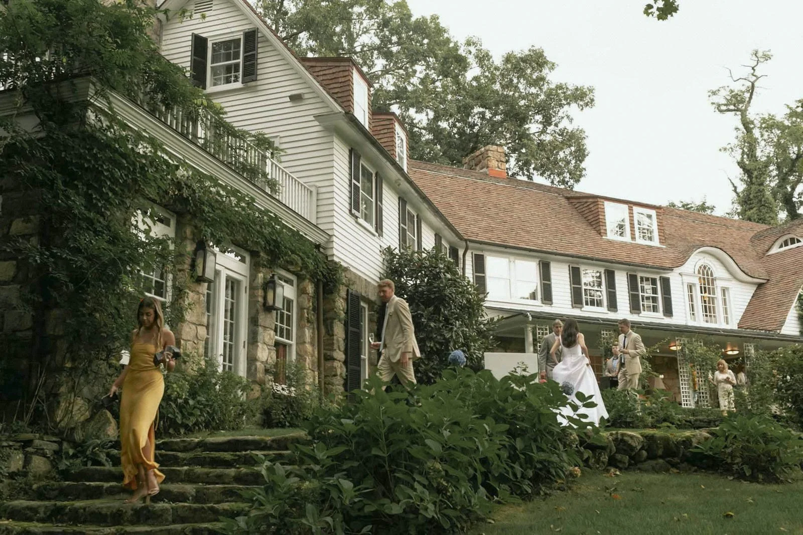 People dressed in formal attire on the steps and patio of a large, elegant house with white siding, stone accents, and black shutters, surrounded by trees and greenery during daytime.