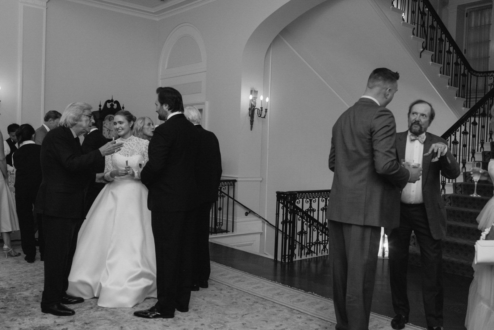 People socializing at a formal event, including a bride in a wedding gown and several men in suits, inside a decorated hall.