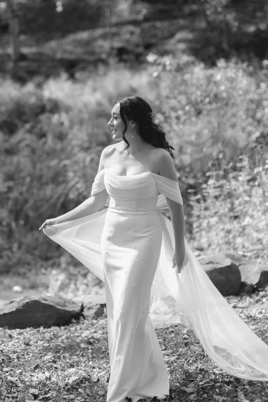A woman in a wedding dress standing outdoors in a natural setting, smiling and holding the edge of her dress.