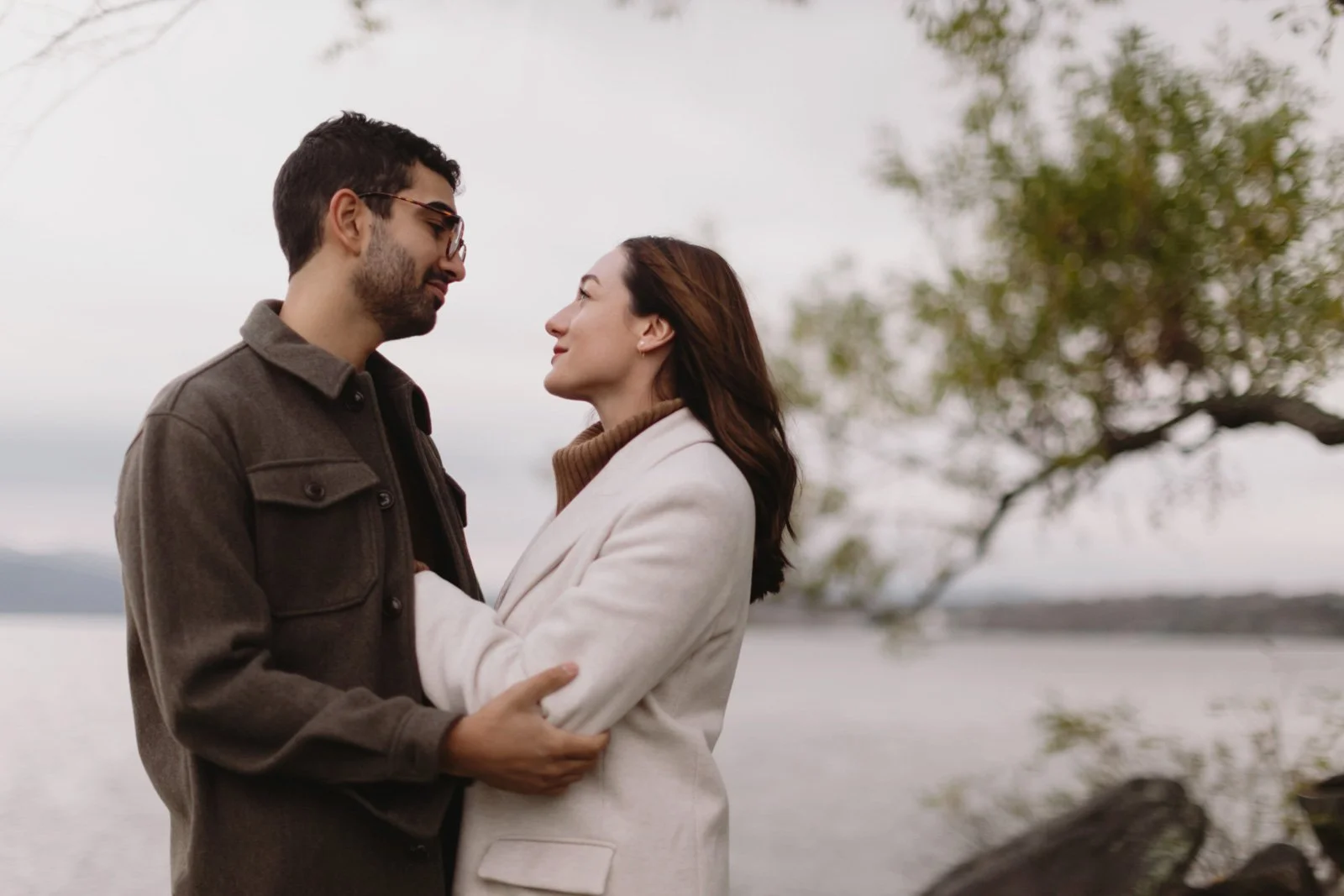 A couple standing close to each other outdoors near a lake, gazing into each other's eyes with a tree and cloudy sky in the background.