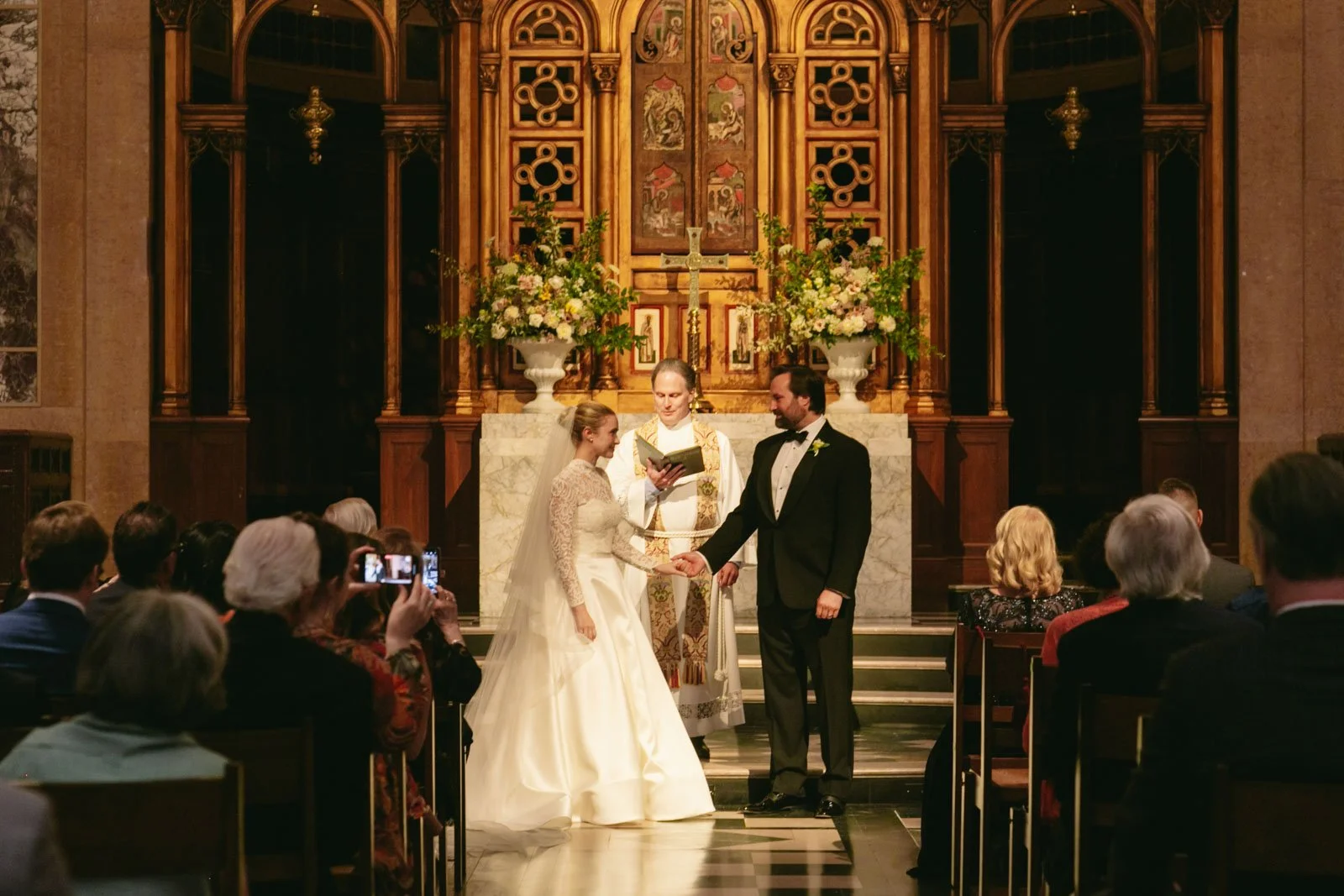 A couple getting married in a church, holding hands in front of an officiant. The bride wears a white lace wedding gown and the groom a black tuxedo. Guests are seated facing them, and some are taking photos. Behind the couple is an ornate wooden alt