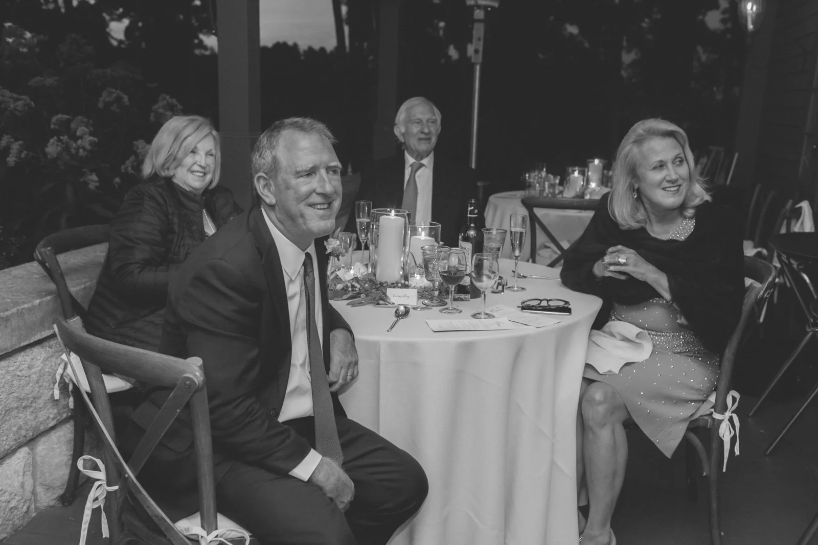 Black and white photo of four people sitting around a decorated table at an evening outdoor celebration, smiling and engaged in conversation.