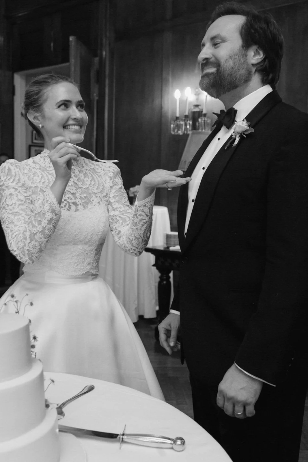 A bride and groom at their wedding, standing in front of a wedding cake. The bride is smiling and holding a knife, preparing to cut the cake. The groom is looking at her, smiling. The scene appears joyful and celebratory, in a room with dark wooden w