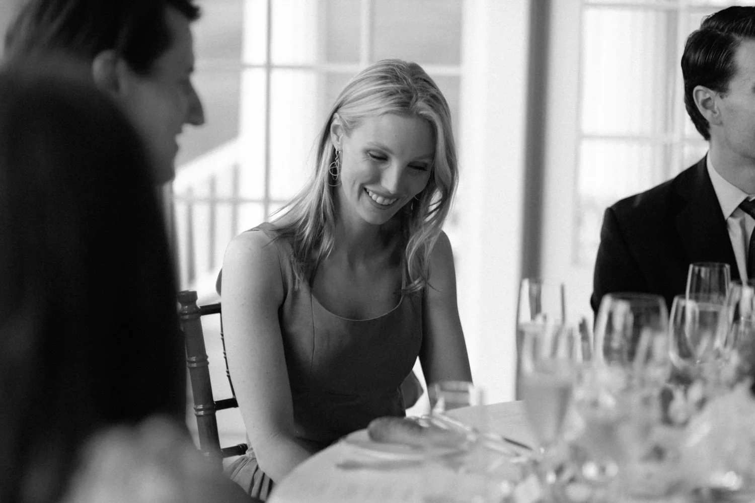 A group of people, including a woman smiling, sitting around a table with wine glasses, in a bright room with large windows.