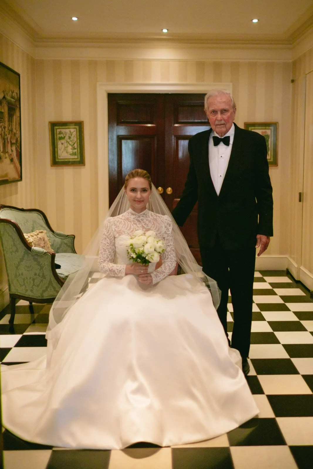 A bride in a white wedding dress and veil sitting on a chair holding a bouquet, and an older man in a black tuxedo standing beside her in a decorated room with framed artwork and checkered flooring.