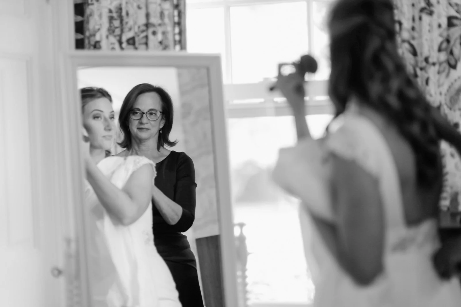 A woman in a wedding dress is getting ready in front of a mirror while another woman, possibly a makeup artist or stylist, is applying makeup or hair. The scene appears to be in a bright room with large windows.