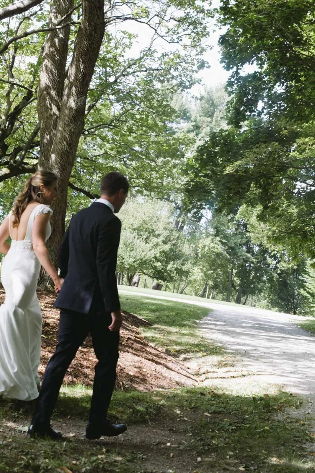 A bride and groom walking hand in hand on a shaded path in a park with green trees and grass.