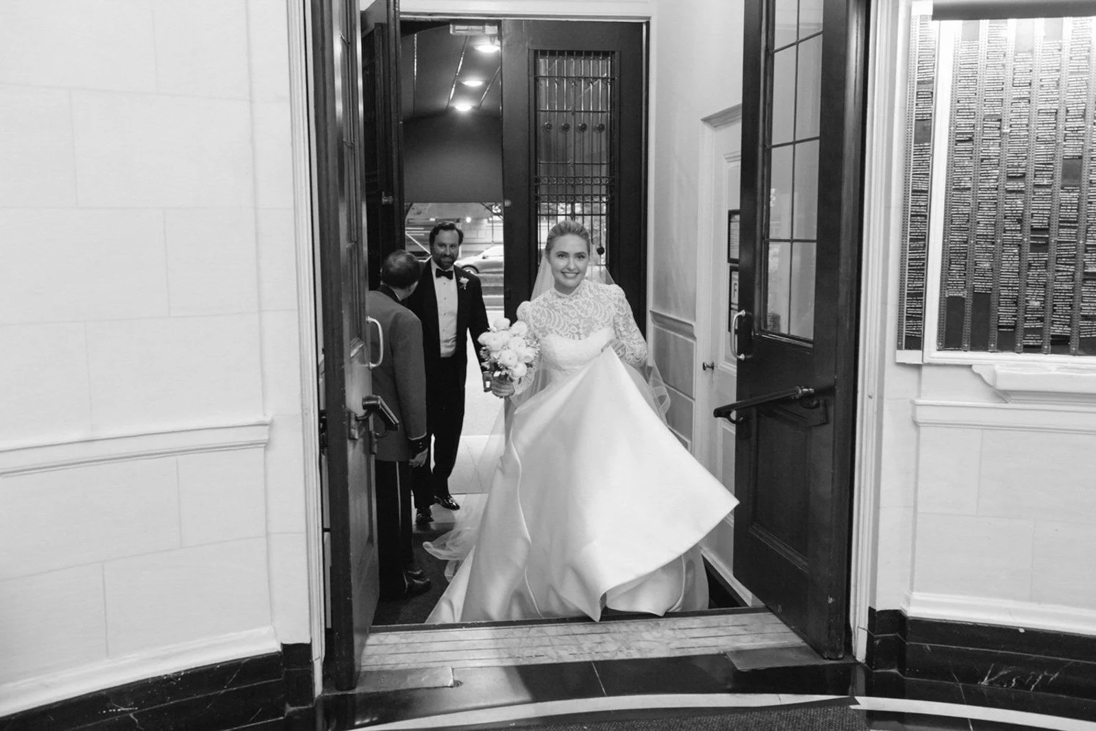 Black and white photo of a bride holding a bouquet and lifting her wedding dress, entering a building with two men in tuxedos inside.