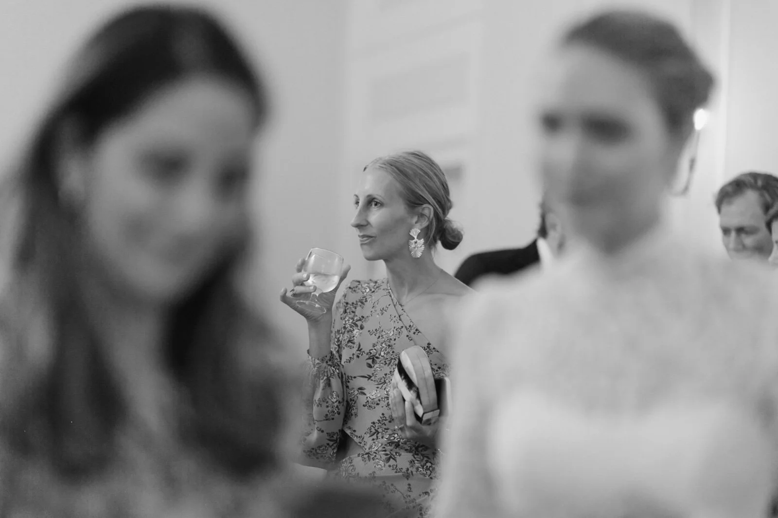 A woman holding a glass of drink, wearing floral dress and large earrings, at a social gathering with blurred faces around her.