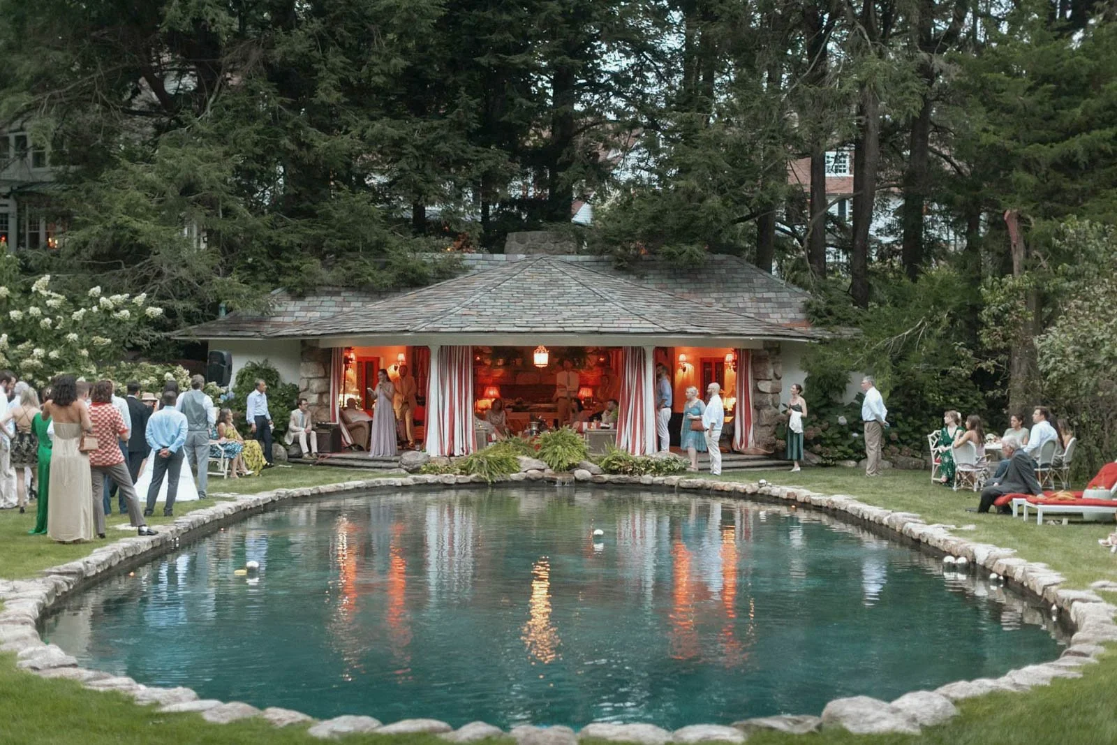 An outdoor gathering with people socializing near a pond and a house with an open patio, surrounded by trees.