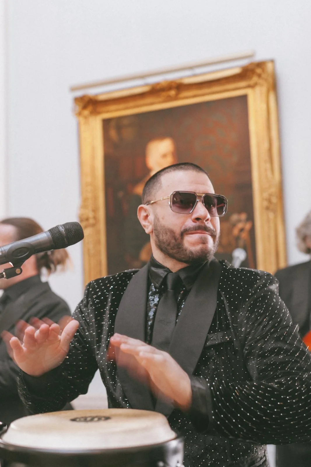 Man with a beard wearing sunglasses and a black blazer with silver dots, clapping during a performance, with a framed painting in the background.