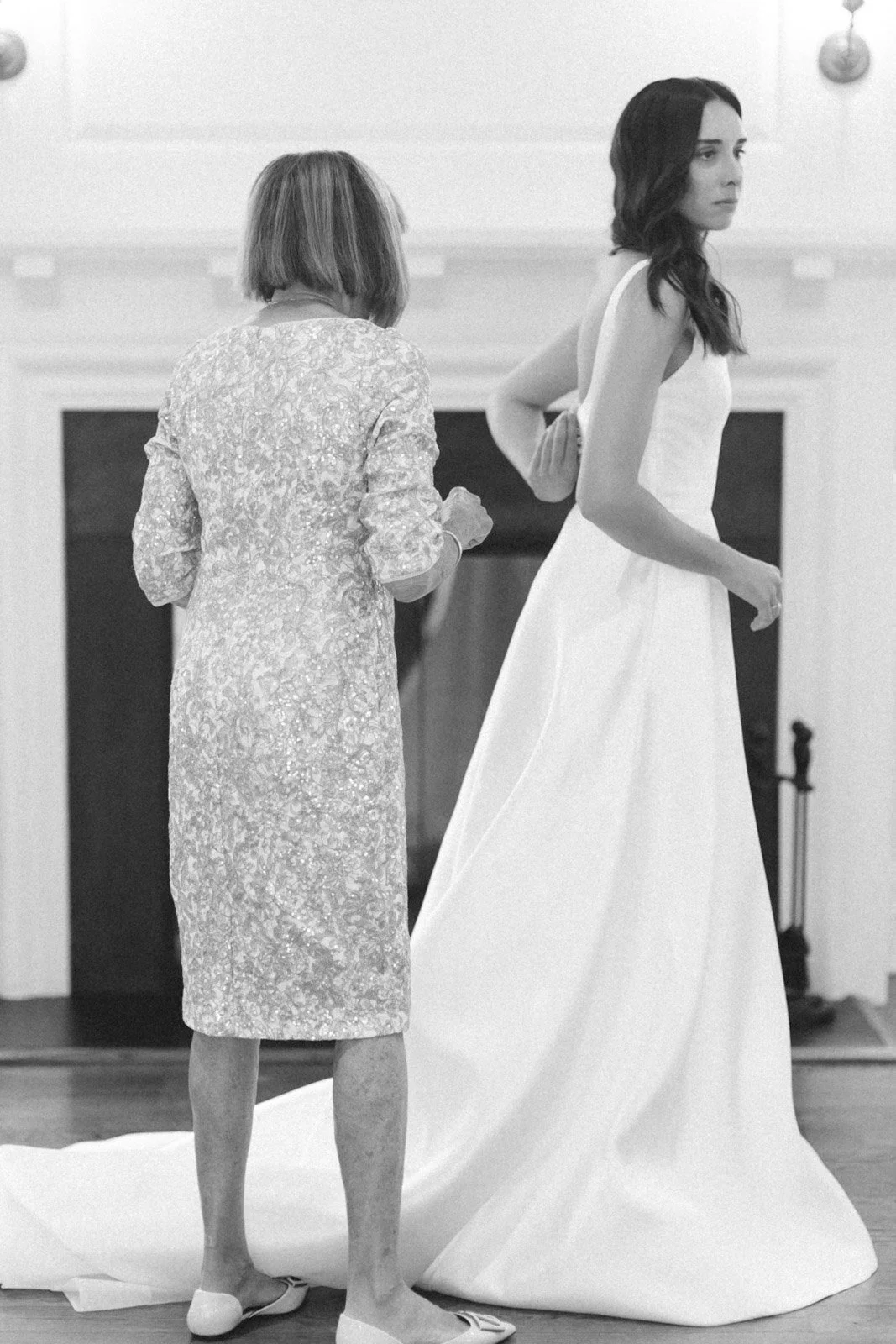 A woman in a wedding dress being assisted by another woman in a patterned coat in a room with a fireplace.