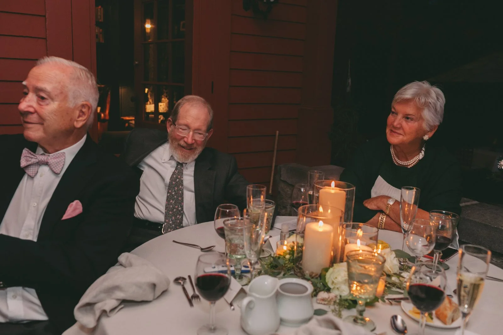 Three elderly people sitting at a round dining table with lit candles, wine glasses, and floral centerpiece, engaging in conversation at a dinner event.