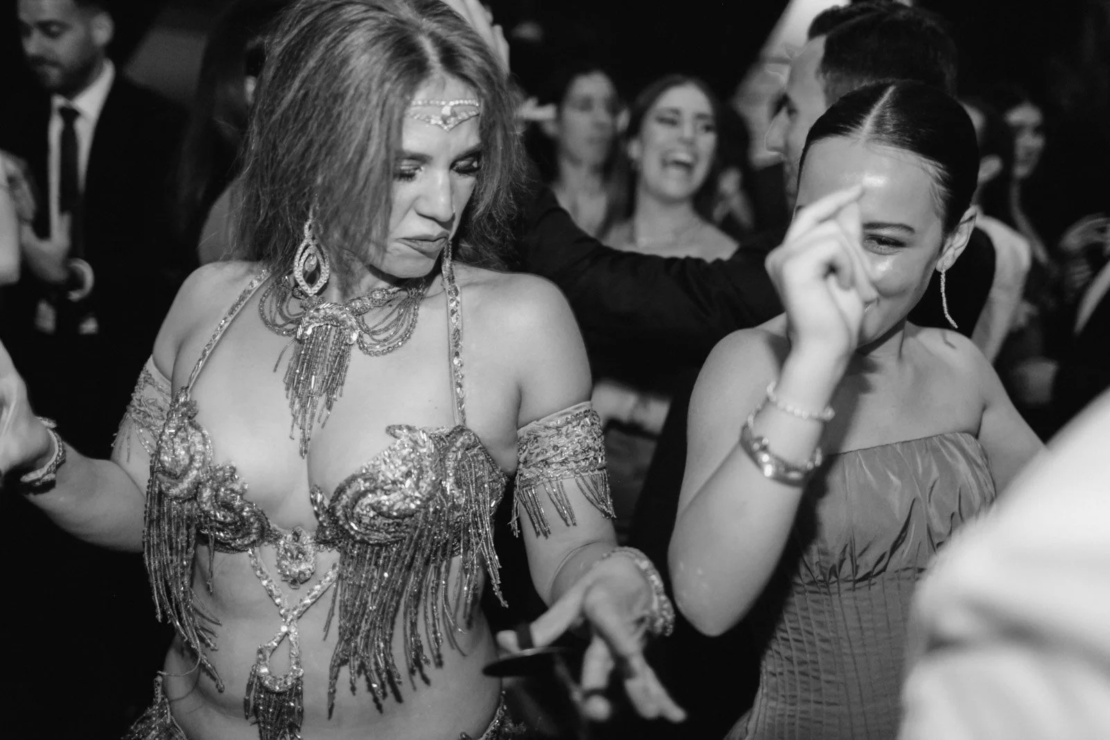 Two women dancing at a social gathering, one dressed in an ornate, beaded costume and the other in a strapless dress, with people dancing in the background.