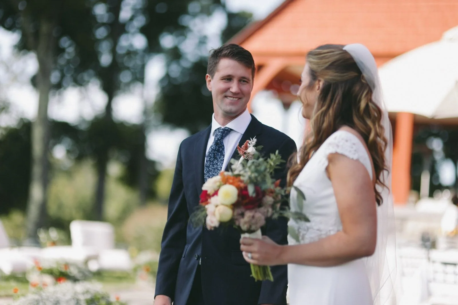 A groom in a dark suit and tie gazes at a bride in a white dress holding a bouquet during a wedding ceremony outdoors.