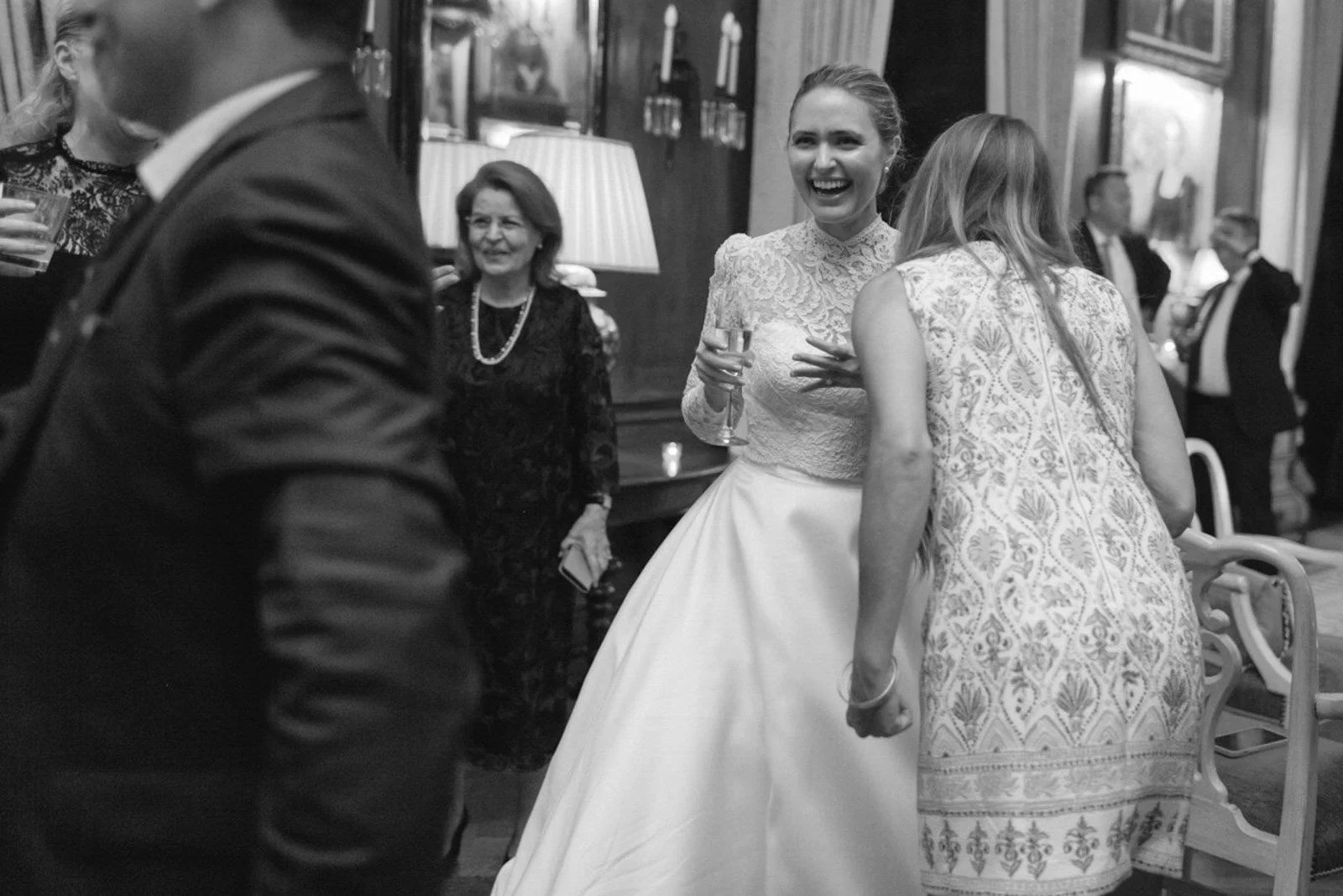 A black and white photo of a wedding reception with guests socializing and laughing indoors. The bride, wearing a lace top and a full skirt, is smiling while holding a glass, and women in patterned dresses are nearby.