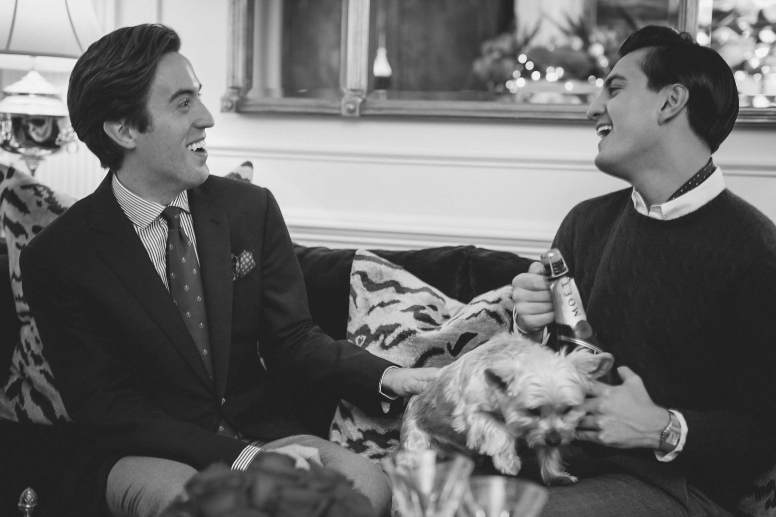 Two men in formal attire laughing, sitting on a sofa with a small dog on the man's lap, holding a bottle, in a decorated room.