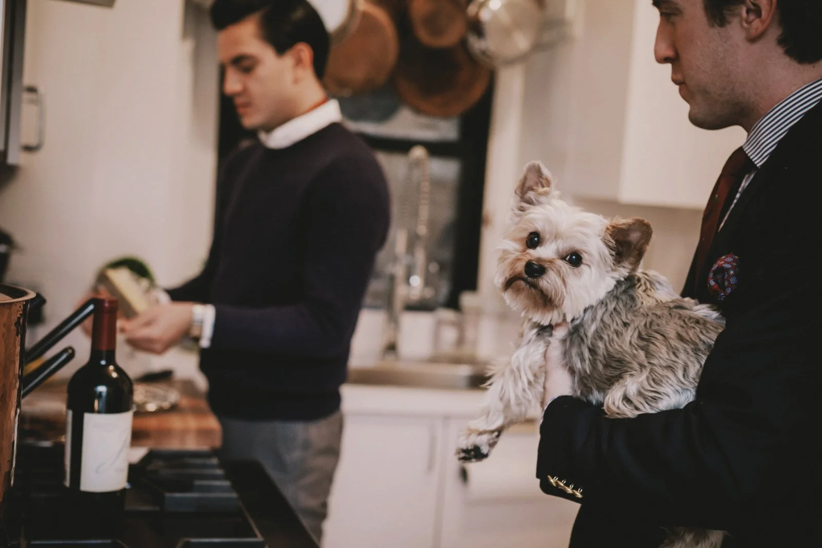 A man holding a small dog inside a kitchen with two people in the background, one of whom is preparing food.
