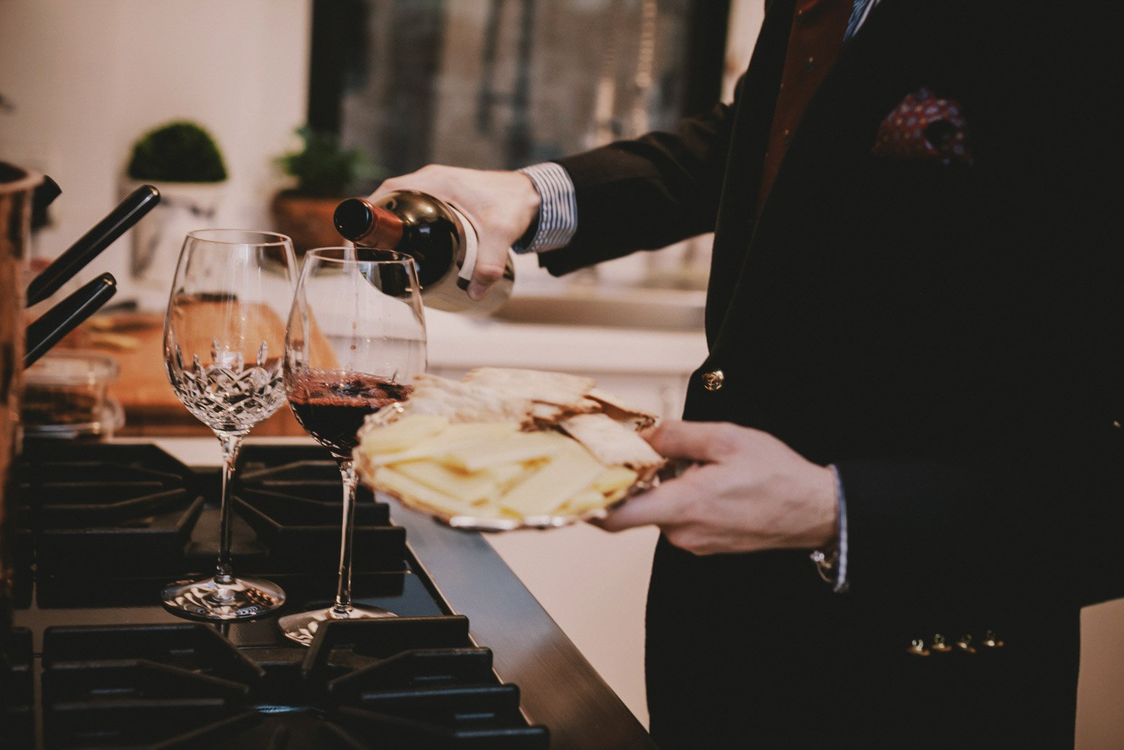 A person in a dark suit pours red wine into a glass while holding a plate of cheese, with a wine glass already filled nearby, on a kitchen countertop.
