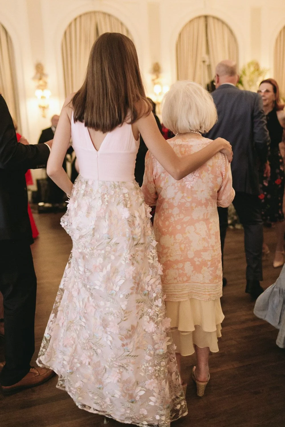 A woman in a white and pink floral dress is walking arm in arm with an older woman in a peach and cream dress at a formal gathering or wedding reception.