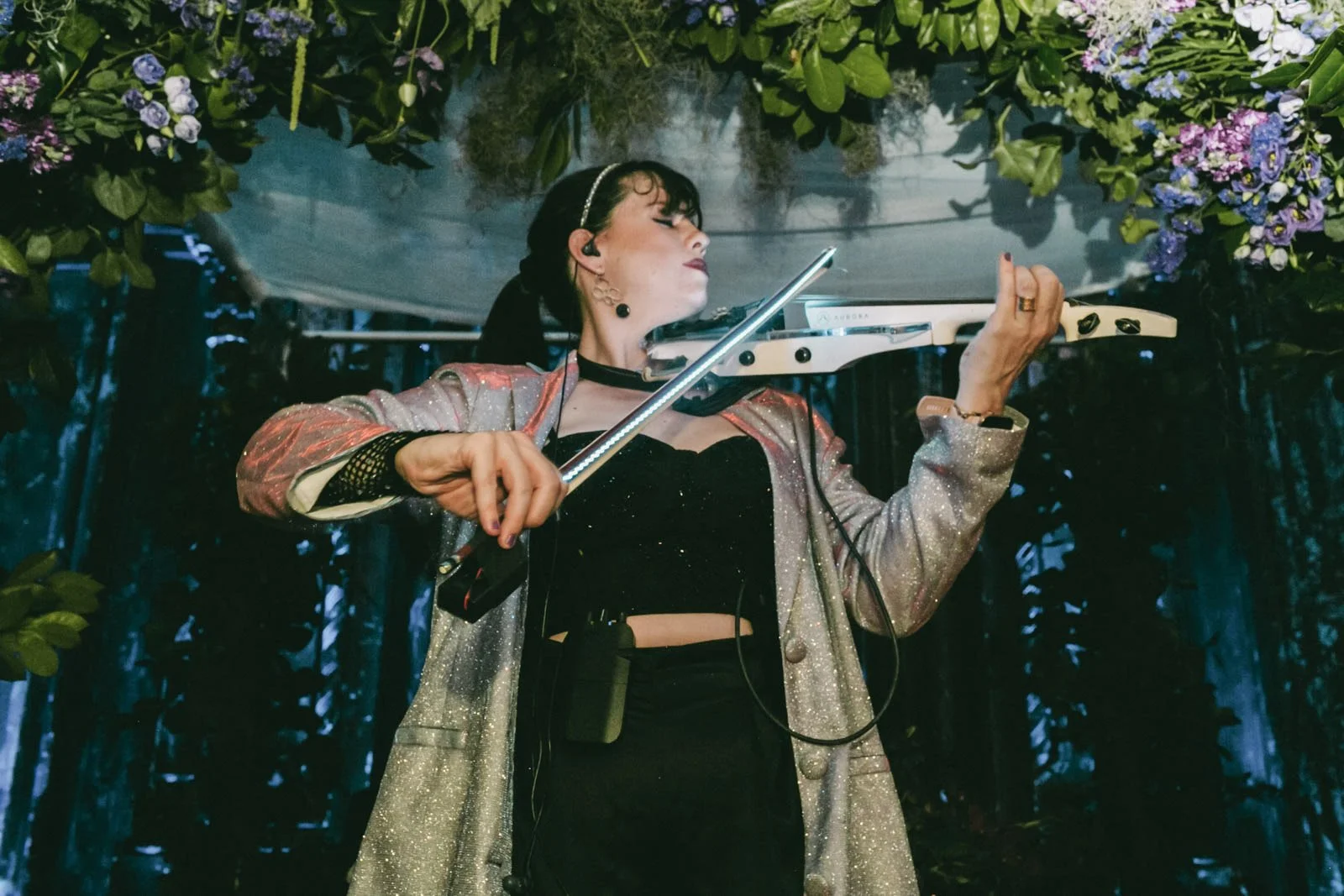 A woman is playing a violin on a stage decorated with green plants and purple flowers.