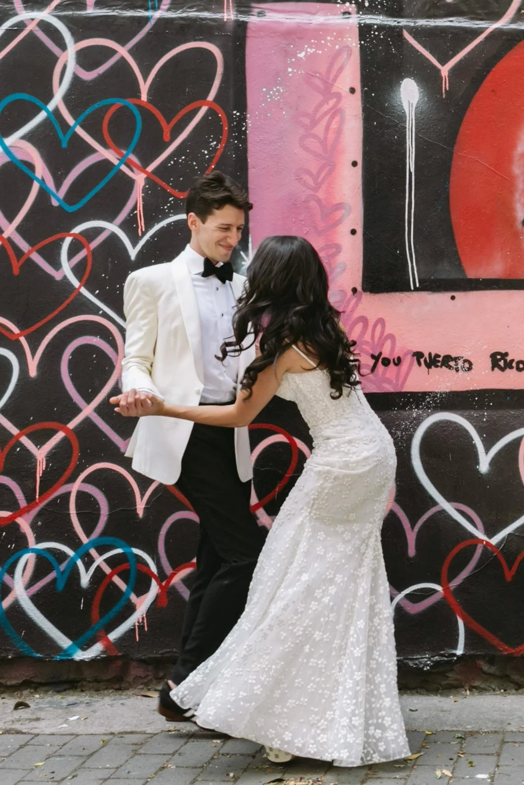 A young man in a tuxedo and a young woman in a wedding dress dancing together outdoors in front of a colorful graffiti wall with hearts and abstract designs.
