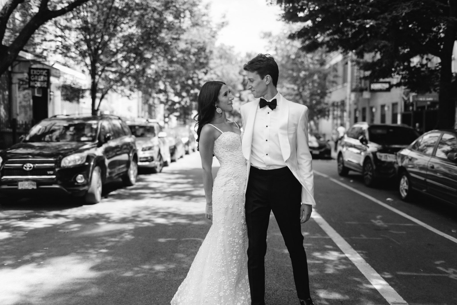 A black and white photo of a bride and groom standing on a city street, looking at each other. The bride is wearing a lace wedding dress and the groom is in a tuxedo with a bow tie.