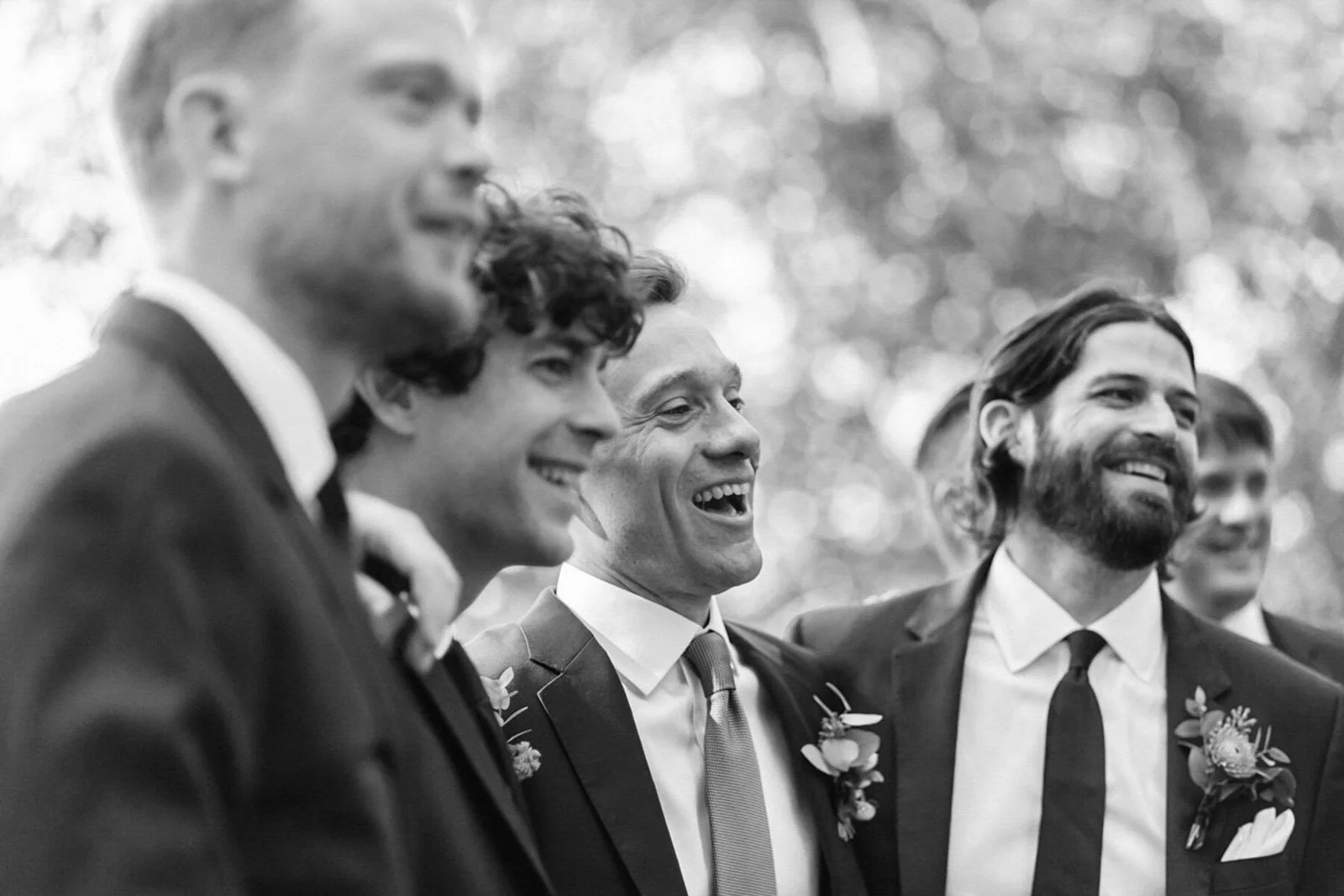 A group of men dressed in suits and ties, smiling and laughing together at an outdoor event with blurred trees in the background.