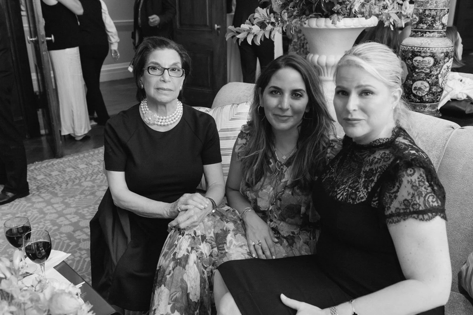 Three women sitting on a sofa at a social event, with two glasses of red wine and a floral centerpiece on a table in front of them.