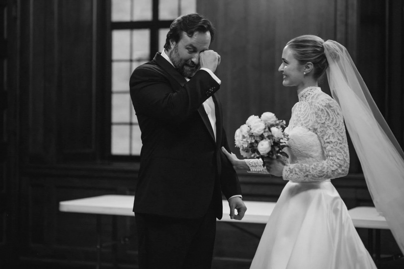 A bride and groom during their wedding ceremony in a dark wooden room with large windows. The groom is emotionally wiping away tears and the bride is smiling while holding a bouquet of flowers.