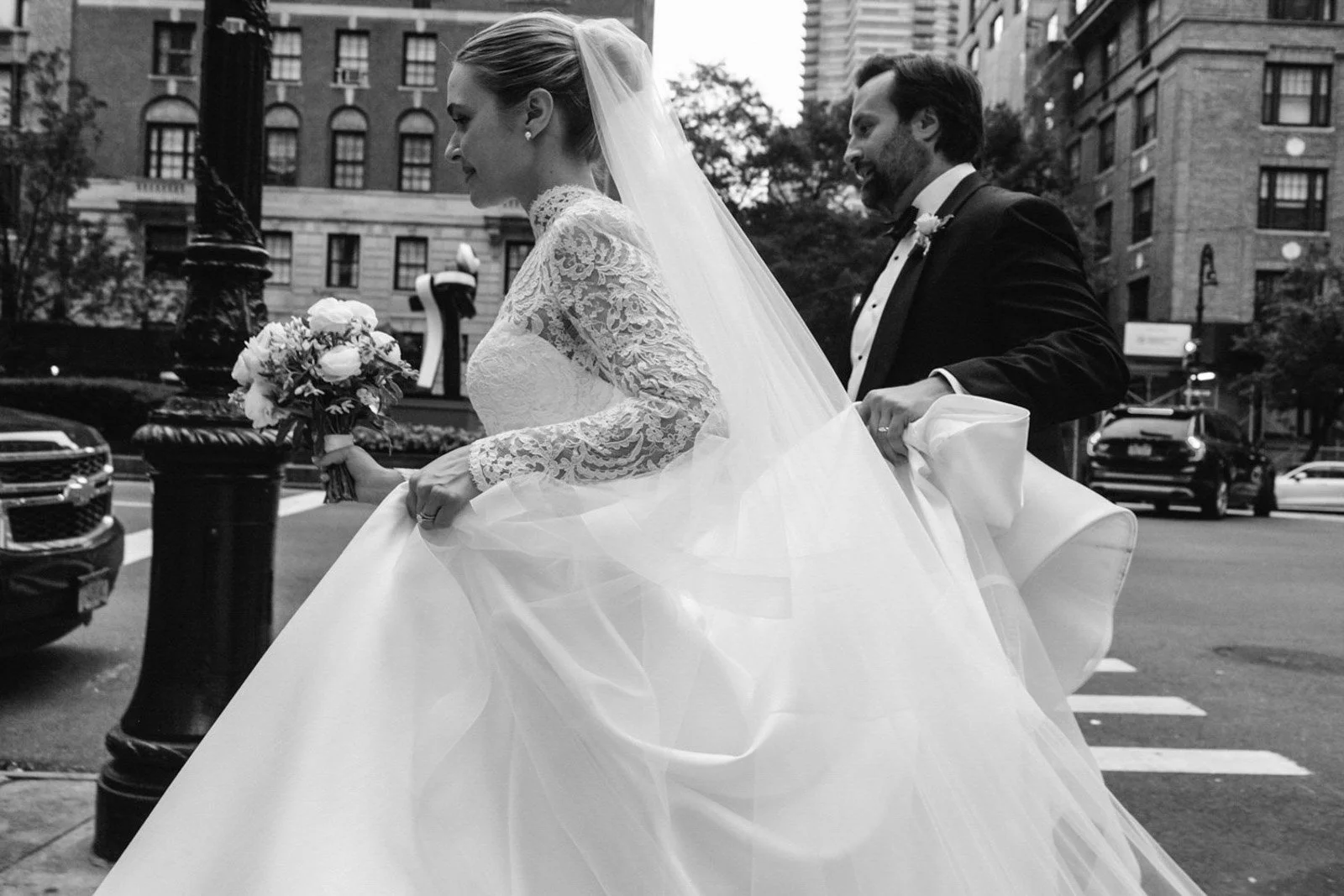 A black and white photo of a bride and groom walking in an urban setting, holding hands. The bride is wearing a long lace wedding dress and veil, holding a small bouquet. The groom is dressed in a tuxedo with a bow tie. Buildings and parked cars are 