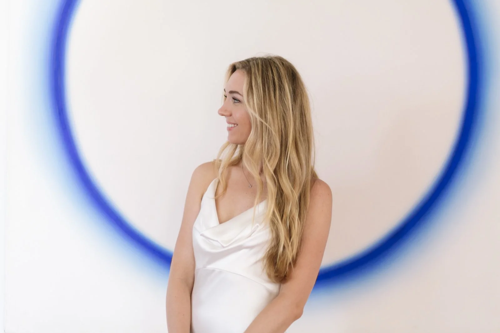 A young woman with long wavy blonde hair, wearing a white satin dress, is smiling and looking to her left. The photo is taken through a circular frame with a blue border, against a plain white background.
