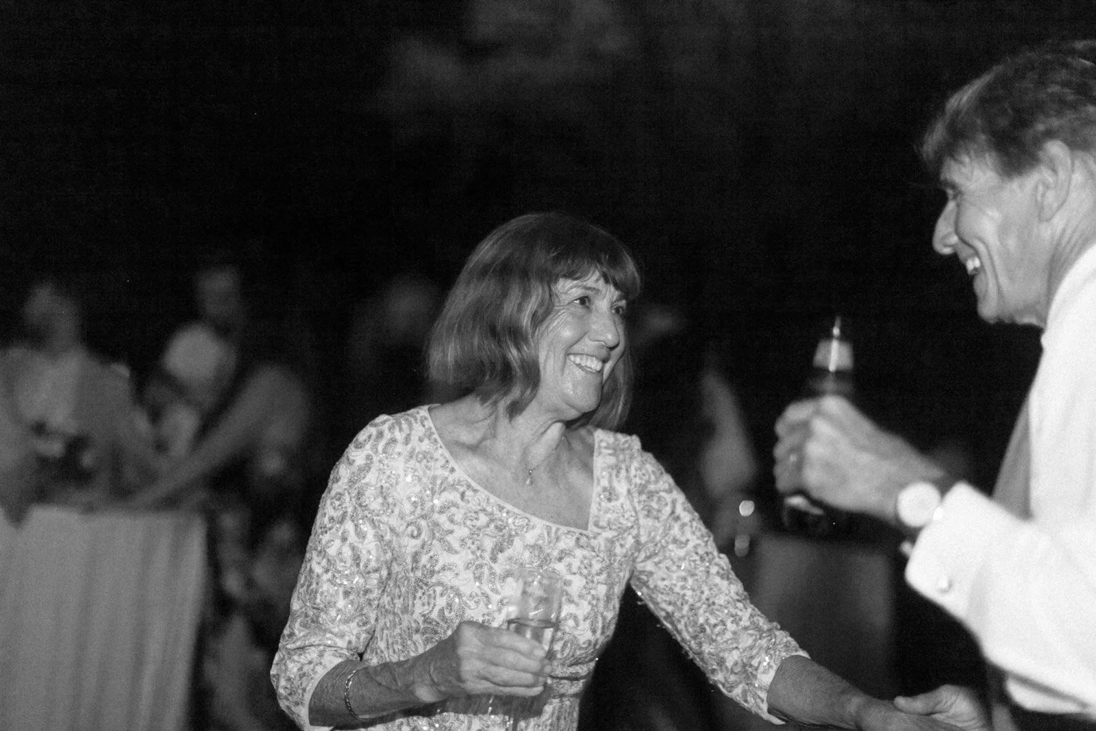 A woman with shoulder-length hair smiling and holding a drink, dancing and interacting with a man holding a beer bottle at a social gathering.