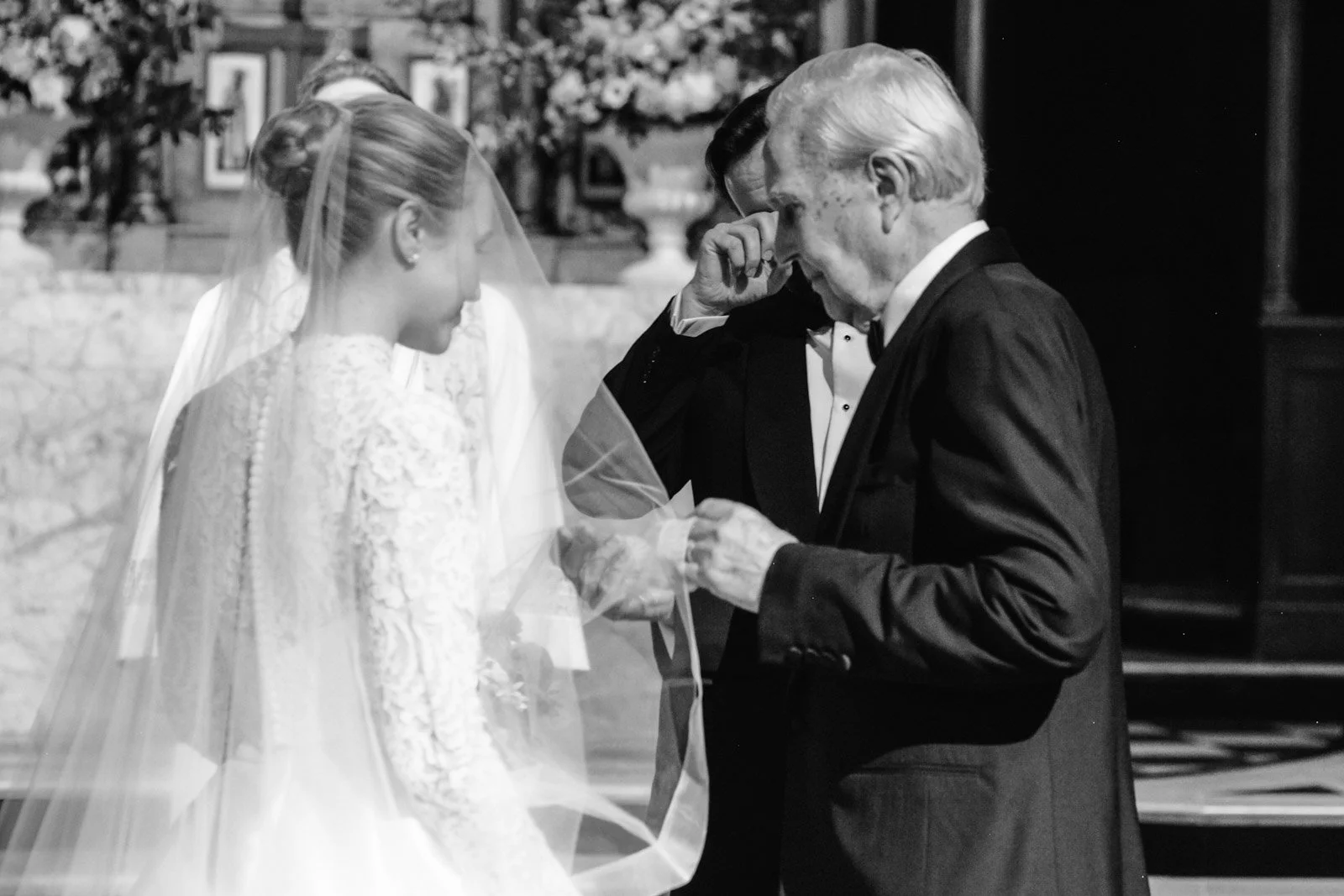 A bride and an older man, possibly her father, at a wedding, sharing a tender moment as he wipes his eye, with another man in the background.