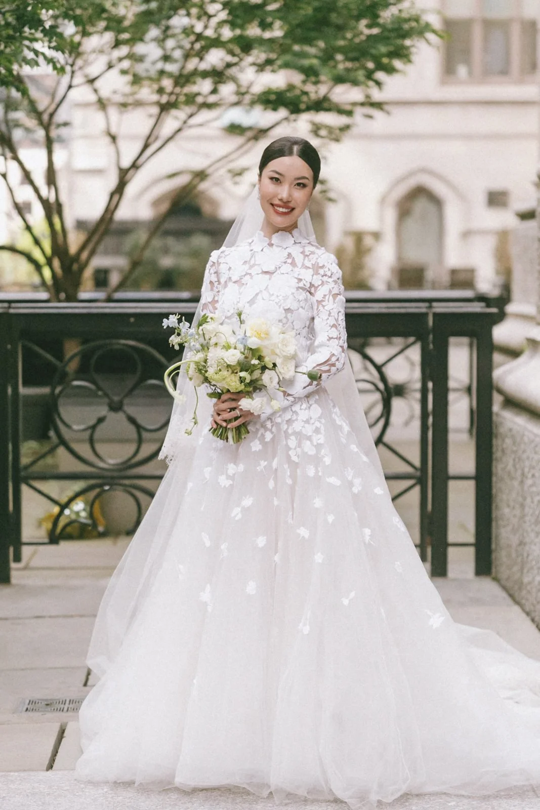 A bride in a white lace wedding dress holding a bouquet of white flowers outside in front of a black railing and a historic building with arched windows, trees, and stone details.