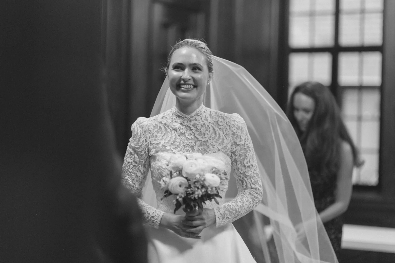 A bride in a lace wedding dress holding a bouquet of flowers, smiling, in a room with large windows, with a bridesmaid in the background.