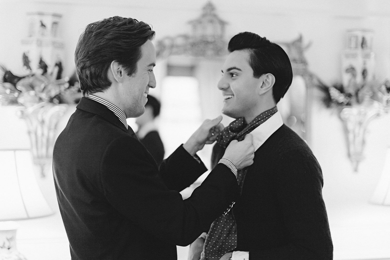 Two men, smiling, are adjusting each other's neckties at what appears to be a formal event or gathering, with a blurred background of a decorated room.