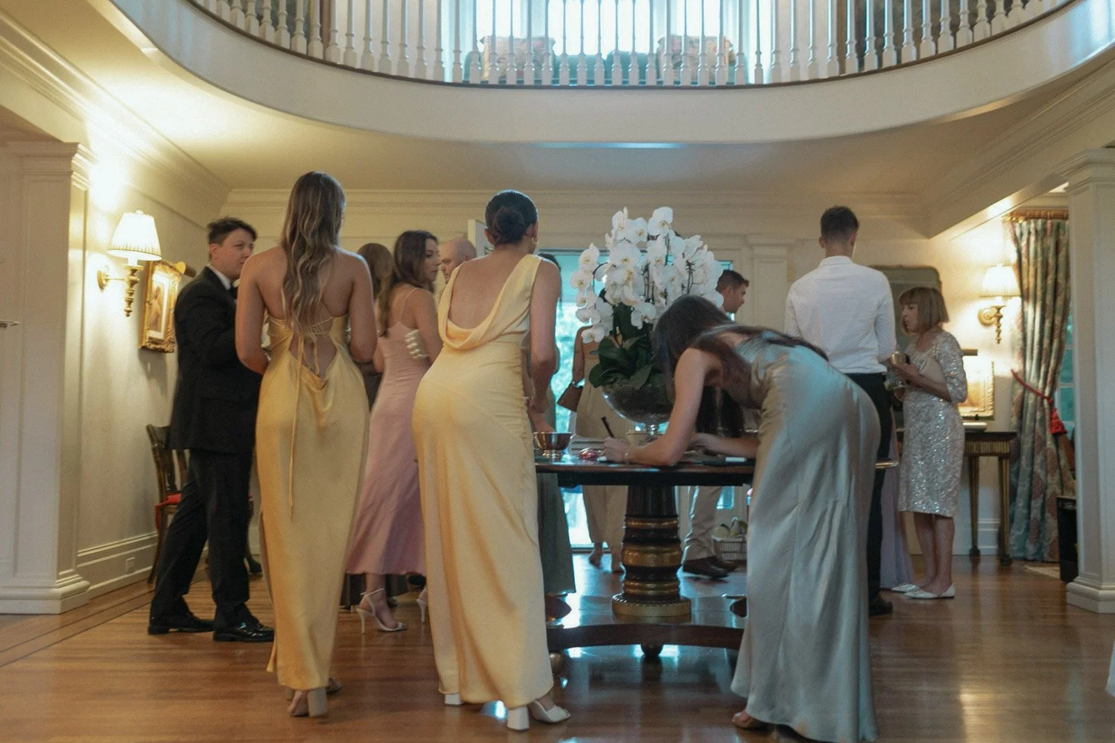 A group of people at a formal event in a decorated indoor setting, with women in evening gowns and men in suits, gathered around a table with a large flower arrangement.