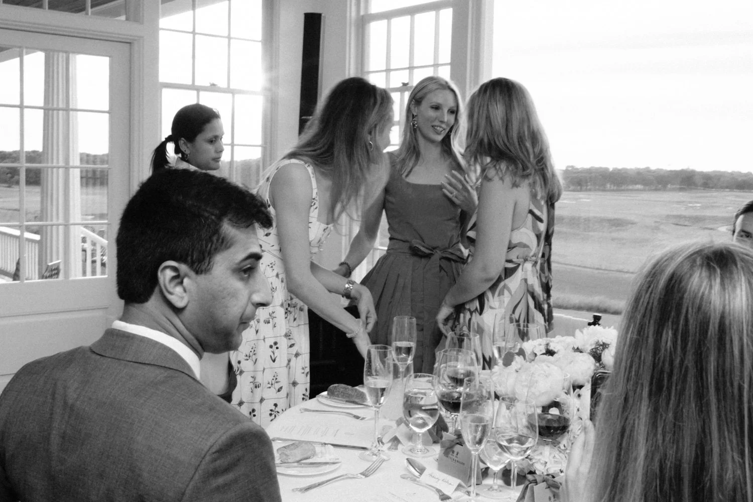 A black and white photo of a group of people at a social gathering, with some standing and talking near a window and others sitting at a table with wine glasses and floral centerpieces.