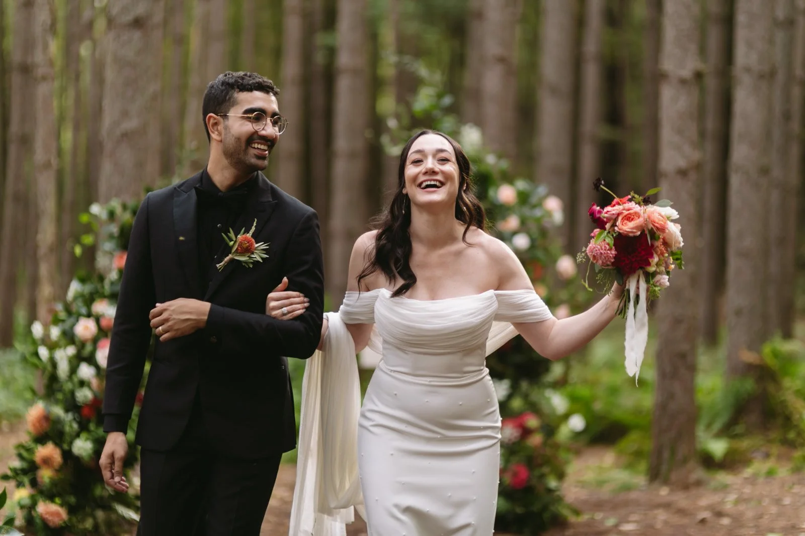 Bride and groom walking arm-in-arm through a forested area, smiling happily, with wedding flowers in the background.