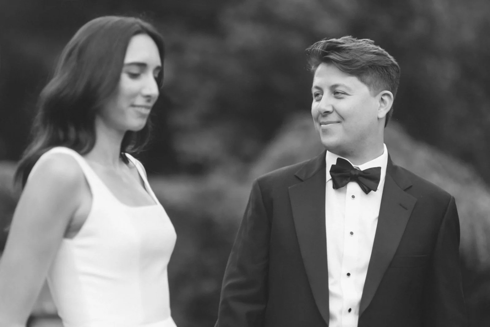 A black and white photo of a woman in a sleeveless dress and a man in a tuxedo with a bow tie, standing outdoors, both looking at each other.