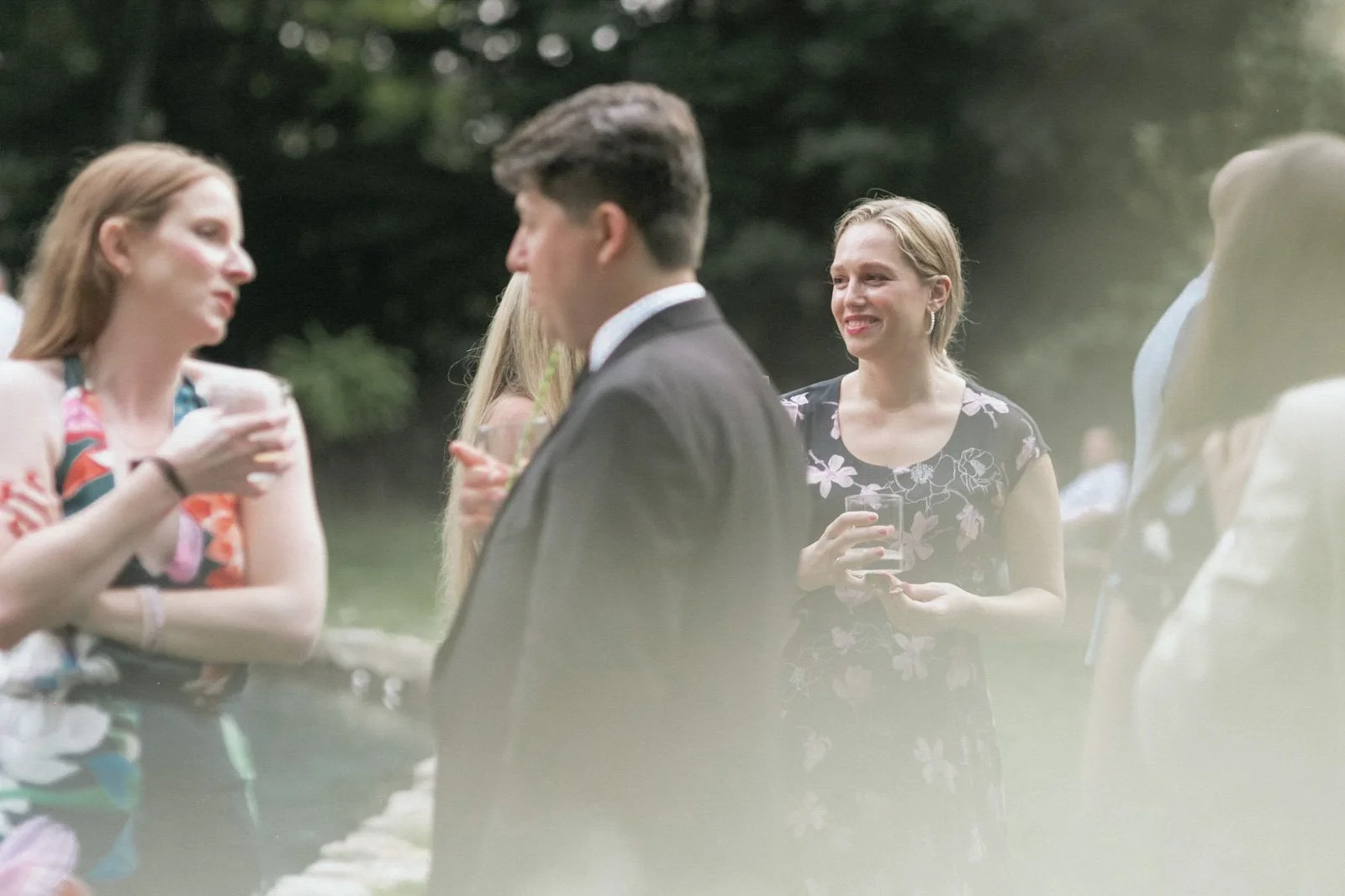 People socializing outdoors during daylight, with a woman in a floral dress holding a drink and smiling.