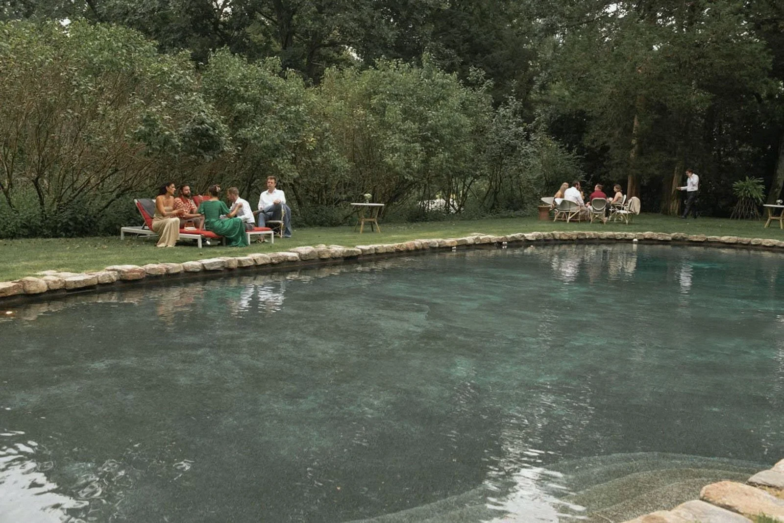 People gathered around tables and on lounge chairs by a swimming pool in a lush garden setting.