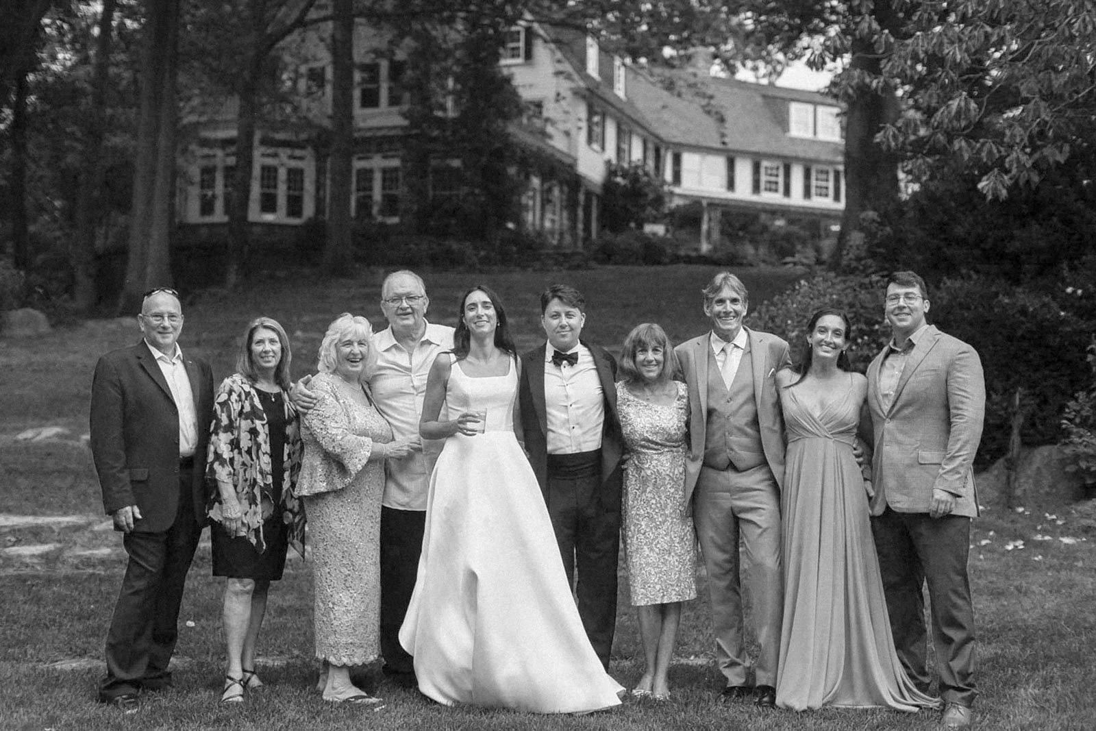 A group of eleven people, dressed formally, standing outdoors on grass with a house and trees in the background, smiling for a photo.