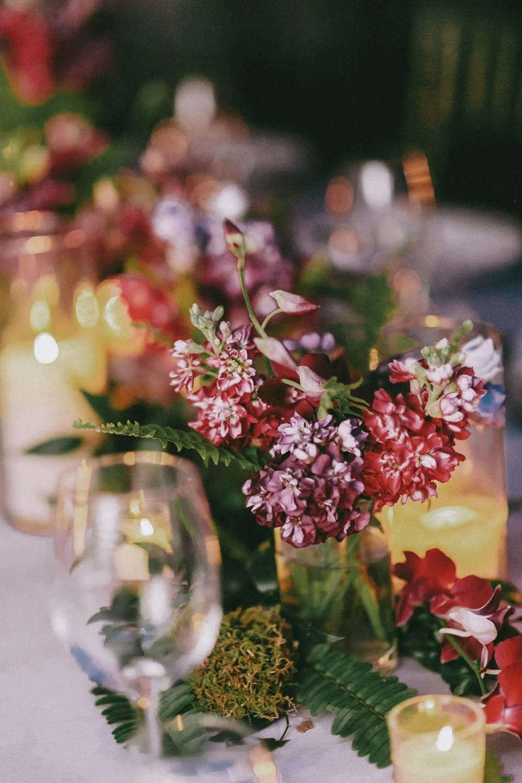Close-up of a floral centerpiece with pink and purple flowers, surrounded by candles and glassware on a decorated table.