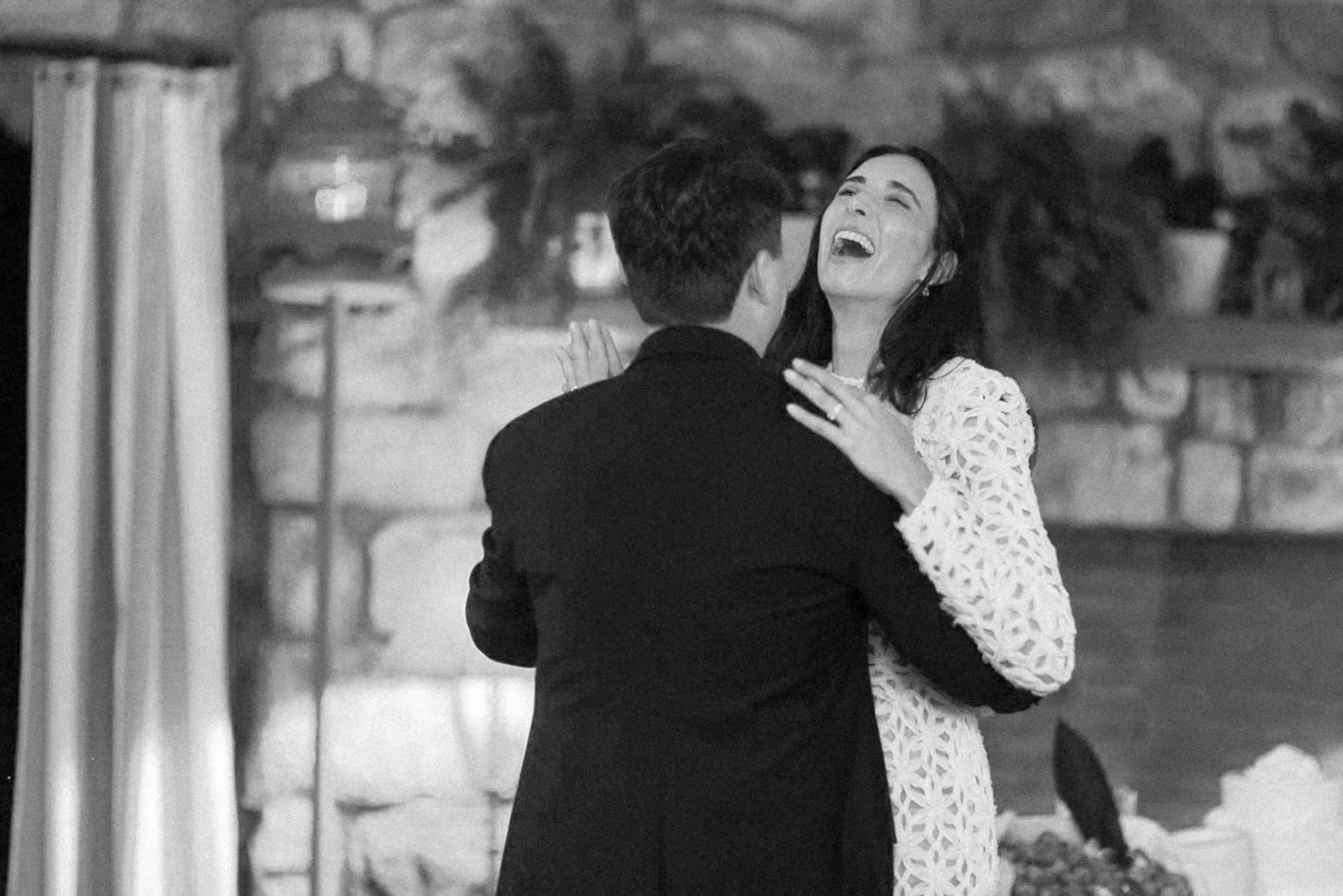 A man in a dark suit and a woman in a white lace dress sharing a joyful dance indoors, with a stone wall and plants in the background in a black and white photograph.
