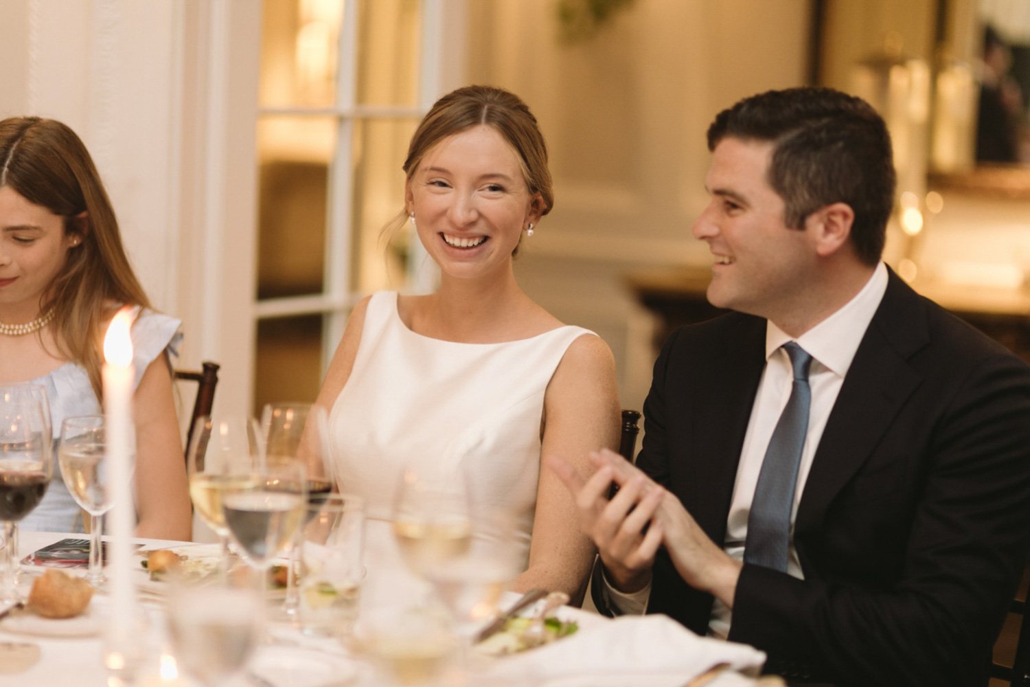 People sitting at a dining table during a celebration, including a woman in a white dress smiling and a man in a suit talking.