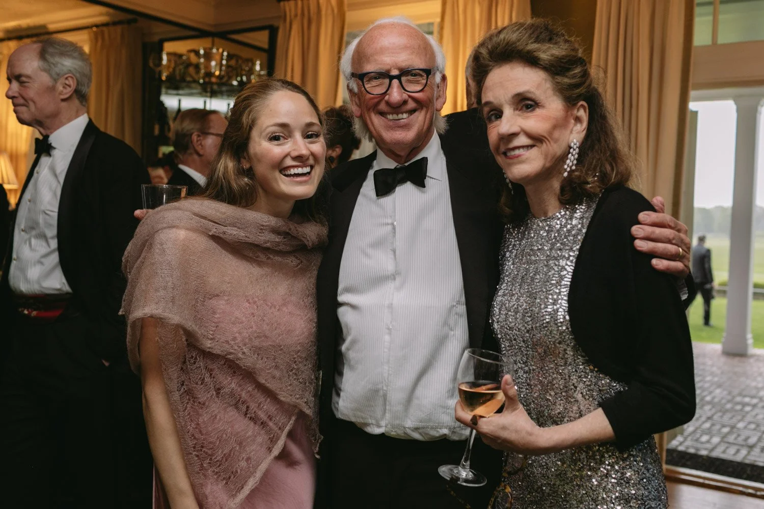 Three smiling people at a formal event, with two women and one man in the center, dressed in evening wear, with the woman on the right holding a glass of wine, standing indoors in a decorated room.