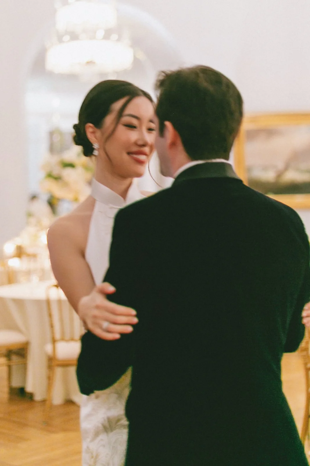 A woman in a white dress and a man in a black suit dancing closely at a formal event, with a smile on her face.