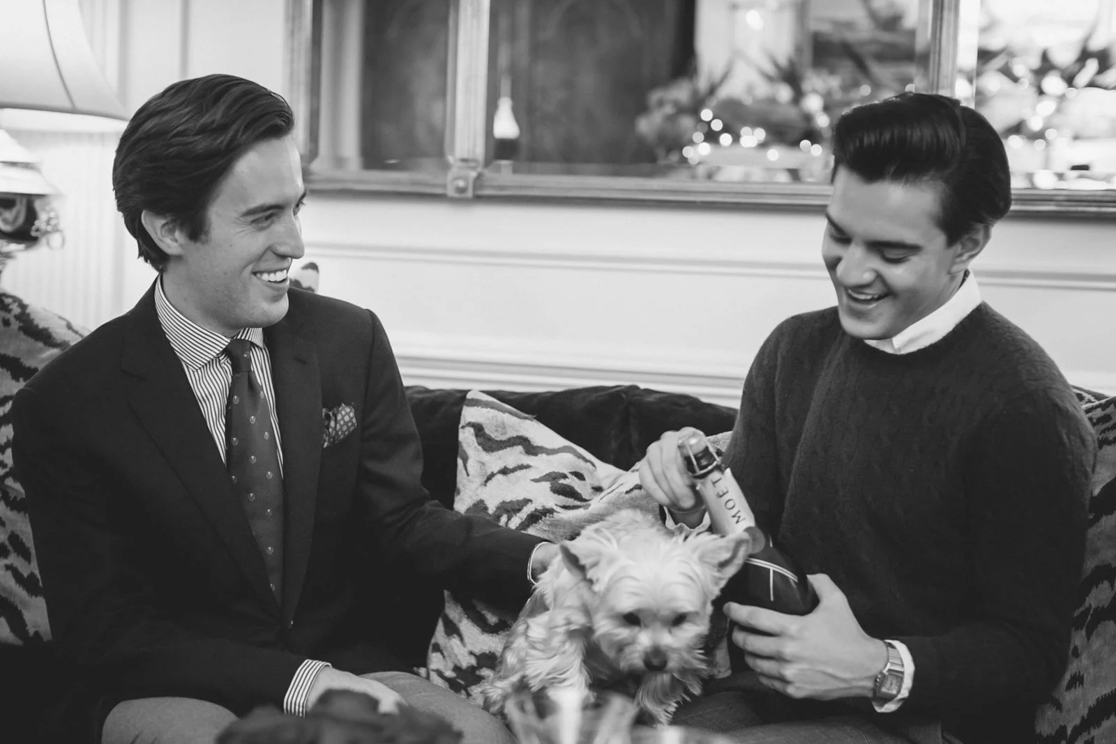 Two men sitting on a sofa with a small dog between them. One man is holding a bottle, and the dog looks at the camera. The men are smiling and seem to be enjoying conversation.